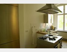 Moderne Küche mit Edelstahlherd und Dunstabzugshaube, weißen Wandfliesen, einem großen Fenster, hellem Holzboden und Arbeitsplatten. Links ein großer Metallkühlschrank, rechts eine Spüle mit Geschirr und Küchengeräten.