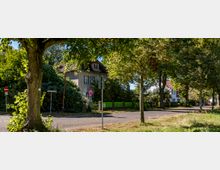 Straßenszene in einer Wohngegend mit Bäumen auf beiden Seiten, einem alten Haus mit Satteldach im Hintergrund und einem grünen Zaun. Verkehrsschilder und ein Gehweg grenzen die Straße ab, während im Hintergrund weitere Häuser sichtbar sind.