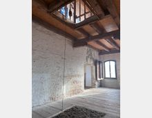 Innenansicht eines rustikalen Raumes mit freiliegenden Holzbalken an der Decke, weiß verputzten und teils freigelegten Ziegelwänden. Der Raum hat ein großes Fenster mit Blick nach draußen und eine schlichte Holzbodenfläche, durchbrochen von einem kleinen Durchgang zu einem angrenzenden Raum.