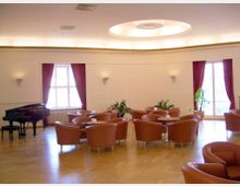 Ein heller Raum mit Holzboden, mehreren runden Tischen und braunen Polstersesseln. Links steht ein schwarzer Flügel, die Fenster sind mit roten Vorhängen dekoriert, und an den Wänden sind schlichte Wandleuchten angebracht.