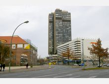 Straßenkreuzung in einer urbanen Umgebung mit einem hohen, modernen Bürogebäude im Hintergrund, einem mehrstöckigen Parkhaus rechts und einem älteren Gebäude mit roten Ziegeldach und Baugerüsten links. Im Vordergrund sind Bürgersteige, Bäume und vereinzelt Passanten zu sehen.