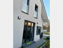 Moderne Hausfassade mit glatter, hellgrauer Putzoberfläche, großen bodentiefen Fenstern und schmalen Glasgeländern an den oberen Fenstern. Im Vordergrund befindet sich ein gepflasterter Weg, im Hintergrund ein Gartenbereich mit einer Rasenfläche und vereinzelten Sitzmöbeln.