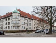Mehrstöckiges Wohnhaus im Altbaustil mit hellen Fassaden, roten Ziegeldächern und dekorativen Erkern. Links im Vordergrund steht ein Straßenschild, im Hintergrund sind geparkte Autos und kahle Bäume an einer gepflasterten Straße zu sehen.