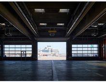 Blick aus einer offenen Halle mit großen Fenstern und Industriedecken auf das Vorfeld eines Flughafens, im Hintergrund ein Kontrollturm und weitere Flughafengebäude. Die Umgebung ist beleuchtet vom Tageslicht.