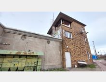 Ein kleines Backsteingebäude mit mehreren Fenstern, einer weißen Tür und einem Flachdach, das an eine Betonwand mit Lüftungsgitter angrenzt. Im Vordergrund befindet sich ein grüner Metallkasten mit Aufschrift, sowie eine weiße Bank und ein schmaler gepflasterter Weg.