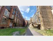 Ein Industriehof mit hohen Backstein- und Betongebäuden auf beiden Seiten, großen rechteckigen Fenstern und teils beschädigten oder verwitterten Fassaden. Im Vordergrund führt ein gepflasterter Weg durch eine grasbewachsene Fläche, während der Himmel teils bewölkt ist.