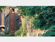 Backsteinbau mit verfallenen Fassadenelementen und Metalltreppe, umgeben von dichtem Grün und Bäumen; im Hintergrund ist ein größeres Industriegebäude zu erkennen.