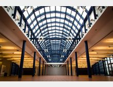 Weitläufiger Innenbereich eines Gebäudes mit einem hohen, gewölbten Glasdach, das von dunkelblauen Stahlträgern gestützt wird. Der Boden ist mit braunen Fliesen bedeckt, und die Wände sind mit Reihen von hängender Beleuchtung ausgestattet.