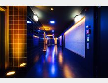Ein schmaler, moderner Flur mit glänzendem dunklem Boden, beleuchtet durch blaues und orangefarbenes LED-Licht. Die Wände bestehen aus schwarzen Kacheln auf der linken Seite und einer hellen, strukturierten Oberfläche auf der rechten Seite; an den Decken befinden sich rechteckige Leuchten.