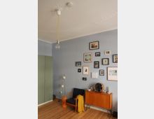 Wohnzimmer mit pastellblauen Wänden, einer Holzkommode mit schlichten schwarzen Beinen, darauf ein Ventilator und dekorative Objekte. Ein Sessel mit blauem Polster und gelber Decke steht daneben, an einer Wand hängen gerahmte Bilder, und die Decke hat eine dekorative Stuckverzierung mit einer Hängeleuchte. Der Holzboden ist hell, und links im Bild ist ein grüner Schrank zu sehen.