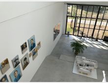 Innenraum einer modernen Galerie mit weißen Wänden, an denen Portraitgemälde hängen. Große deckenhohe Fenster lassen viel natürliches Licht herein; im Raum stehen ein Architekturmodell, eine Zimmerpflanze und mehrere Ventilatoren.