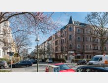 Straßenszene in einem Wohnviertel mit mehrstöckigen Altbauten, darunter ein Eckhaus mit Türmchen und Kletterpflanzenfassade. Im Vordergrund blühende Bäume, parkende Autos und Straßenschilder unter einem klaren, blauen Himmel.