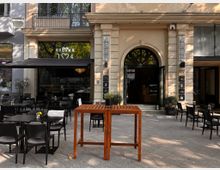 Straßencafé vor einem Gebäude mit heller Fassade und großen Eingangsflügeltüren, flankiert von zwei hohen Säulen. Unter einer schwarzen Markise stehen schwarze Tische und Stühle, einige mit Blumenvasen dekoriert, auf einem gepflasterten Gehweg.