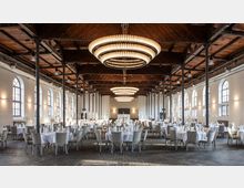 Großer Veranstaltungsraum mit hohen Decken und Holzdach, dekoriert für ein Bankett. Der Raum ist geprägt von großen runden Kronleuchtern, mehreren gedeckten Tischen mit weißen Tischdecken sowie hohen Fenstern mit Rundbögen, die Tageslicht hereinlassen.