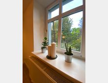 Fensterbank mit drei Dekorationselementen: zwei kleine Topfpflanzen und drei weiße Kerzen auf einem runden Tablett. Im Hintergrund ist ein großes Fenster mit Blick auf einen grünen Garten zu sehen, darunter befindet sich ein Heizkörper.