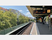 Ein oberirdischer U-Bahnhof mit überdachtem Bahnsteig, Gleisen und einer digitalen Anzeigetafel. Im Hintergrund sind grüne Bäume und Wohnhäuser unter einem blauen Himmel sichtbar.