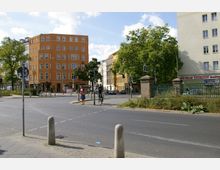 Straßenszene in einer Stadt mit einem markanten, orangefarbenen Wohn- und Geschäftshaus im Mittelpunkt. Im Vordergrund eine Kreuzung mit Fußgängern, einem Radfahrer und Grünflächen, umgeben von weiteren Gebäuden und Bäumen.