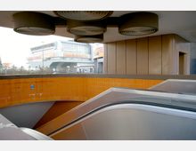 Ein U-Bahn-Zugang mit einer Rolltreppe, umgeben von orangefarbenen Fliesenwänden und einer Betonüberdachung mit runden Lüftungselementen. Im Hintergrund ist ein modernes Gebäude mit einer Glasfassade und Werbebannern sichtbar.