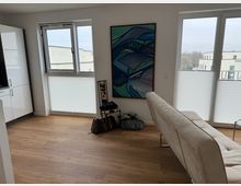 Helles Wohnzimmer mit Holzboden, großen Fenstern mit weißen Plissees und moderner Einrichtung. Ein beige Sofa, ein abstraktes Gemälde an der Wand und ein Teil einer weißen Küchenschrankfront sind sichtbar.