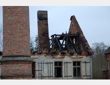 Teilweise abgebranntes Gebäude aus Ziegelstein und Fachwerk mit stark beschädigtem Dach und schwarzen verkohlten Balken. Im Vordergrund steht ein hoher, runder Backsteinkamin.