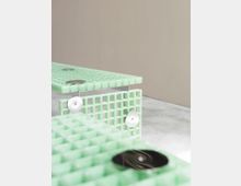 Grüne, gitterartige Möbelstücke mit rechteckigen Formen stehen auf einem glatten, grauen Boden vor einer beigefarbenen Wand. Die horizontale Oberfläche der Möbel ist mit runden Metallelementen versehen.