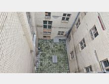 Blick in einen engen, rechteckigen Innenhof mit Fassaden aus hellen Ziegelsteinen. Der Boden ist mit vermoosten quadratischen Fliesen bedeckt, und die umliegenden Gebäude zeigen alte, teils beschädigte Fenster.