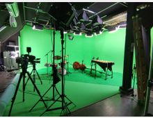 Aufnahme eines Studios mit grünem Greenscreen-Hintergrund, mehreren professionellen Kameras auf Stativen, Beleuchtungsgeräten und Musikinstrumenten wie Schlagzeug, Kontrabass und Vibraphon.