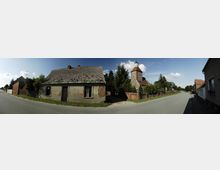 Straßenszene in einem Dorf mit einem alten, leicht verfallenen Haus links im Bild, einer gepflasterten Straße und einer historischen Kirche aus Stein mit Turm und rotem Ziegeldach im Hintergrund, umgeben von Bäumen und einer niedrigen Ziegelmauer. Moderner Asphalt verläuft zwischen den Gebäuden unter blauem Himmel mit wenigen Wolken.