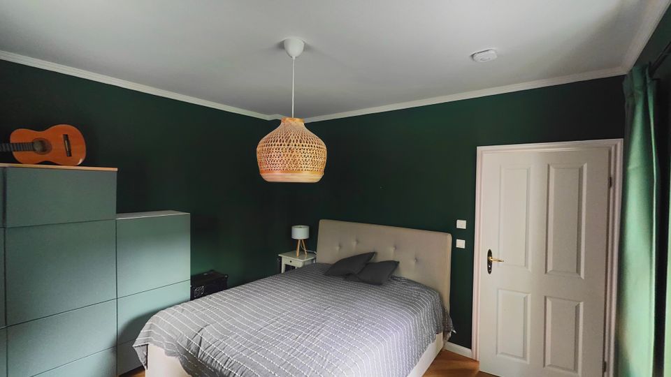 Schlafzimmer mit dunkelgrünen Wänden, Parkett im Fischgrätmuster und einem Doppelbett mit gepolstertem Kopfteil sowie grauer Tagesdecke. An der Decke hängt eine geflochtene Pendelleuchte; neben dem Bett stehen ein Nachttisch, links eine Kommode, rechts eine weiße Tür und ein Fenster mit grünen Vorhängen.