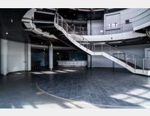 Großer, moderner Innenraum mit dunklem Bodenbelag und geschwungener Metalltreppe, die zu einer umlaufenden Galerie führt. Der Raum verfügt über helle Wände, runde Säulen und eine zentrale Thekenfläche.