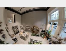 Innenansicht eines kleinen Ladens mit dekorativen Produkten, Pflanzen und Keramikartikeln. Der Raum hat große Fensterfronten, helle Wände und Regale mit Waren sowie einen zentralen Tisch mit Büchern und Dekoobjekten.