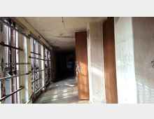 Langer, leerer Flur in einem heruntergekommenen Gebäude mit rissigen Wänden und Decke sowie Betonboden. Links verläuft eine Reihe hoher Fenster mit Metallgittern und Jalousien, rechts stehen mehrere braune Türen entlang des Gangs.