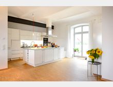 Moderne Wohnküche mit weißer Einbauküche, zentraler Kochinsel und offenen Regalen. Der Raum hat helle Holzböden, hohe Decken, ein großes Fenster mit Vorhängen und eine Vase mit Sonnenblumen auf einem Beistelltisch.