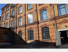 Fabrikgebäude aus rotem und gelbem Backstein mit hohen, bogigen Fenstern, die teilweise vergittert sind. Metalltreppen verlaufen an der Fassade, und eine offene Tür führt ins Gebäudeinnere; der Hof liegt im Schatten.