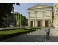 Das Bild zeigt die Fassade des Maxim-Gorki-Theaters in Berlin, ein klassizistisches Gebäude mit hellbeigem Putz, großen Eingangstüren aus Holz und Säulenverzierungen. Im Vordergrund befindet sich ein gepflasterter Weg, sowie Grünflächen mit niedrigen Hecken, und rechts sind geparkte Fahrräder zu sehen.