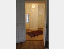 Blick durch geöffnete doppelflügelige weiße Holztüren in einen Raum mit Holzboden, einem roten Orientteppich, weiß gestrichenen Wänden und einem kleinen Kronleuchter. Im Hintergrund sind eine Milchglastür und eine weitere Holztür zu sehen.