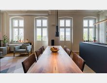 Wohnbereich mit Esszimmer und moderner Einrichtung. Ein langer Holztisch mit Stühlen steht im Vordergrund, dahinter sind große Fenster mit Blick ins Freie, daneben befindet sich eine schlichte Küche mit einer dunklen Arbeitsplatte.