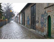 Nasse, kopfsteingepflasterte Gasse mit alten Backsteingebäuden links und rechts; einige der Wände sind mit Graffiti bedeckt, und am Ende der Straße sind weitere Häuser und ein Baum zu sehen. Links ist ein schmiedeeiserner Zaun erkennbar, dahinter Bäume und spärliche Weihnachtsdekoration.