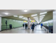 U-Bahn-Station mit grün gefliesten Wänden, heller Beleuchtung und einem Bahnsteig auf der rechten Seite. Mehrere Personen bewegen sich oder warten, und es sind Hinweisschilder sowie Sitzbänke sichtbar.