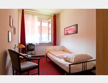 Ein schlicht eingerichtetes Einzelzimmer mit einem Einzelbett, rotem Teppich, einem kleinen Fernseher auf einem Metalltisch und einem Stuhl. Im Hintergrund befindet sich ein weit geöffnetes Fenster mit roten Vorhängen und Blick nach draußen.