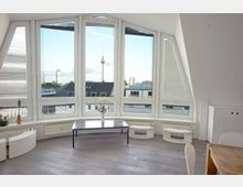 Wohnraum mit großen, dreieckigen und rechteckigen Fenstern, die einen Blick auf die Stadtsilhouette mit dem Berliner Fernsehturm bieten. Innen befinden sich minimalistische Möbel, darunter ein rechteckiger Couchtisch, weiße Schränke und Dekoration mit Pflanzen.