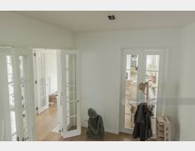 Heller Innenraum mit Holzboden, weißen Wänden und mehreren verglasten Doppeltüren, die in angrenzende Räume führen. Im Vordergrund stehen eine Skulptur, ein Garderobenständer mit Hüten und Jacken sowie ein Kartonregal.