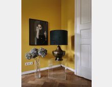 Ecke eines Raumes mit gelber Wand, einem gerahmten Porträtbild, einer durchsichtigen Vase mit getrockneten Hortensien, einer Tischlampe mit dunklem Schirm auf einem gläsernen Beistelltisch und einer weißen Tür mit klassischen Kassettenelementen. Der Boden besteht aus Fischgrätparkett.