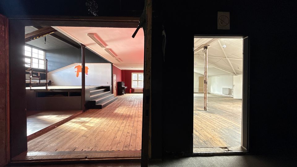 Blick durch zwei Öffnungen in eine ehemalige Veranstaltungshalle mit breitem Holzdielenboden, einer kleinen Bühne mit Stufen und großen Fenstern an der Stirnseite. Rechts ist ein angrenzender leerer Raum mit sichtbaren Stützpfeilern und hoher, schräger Decke zu sehen.
