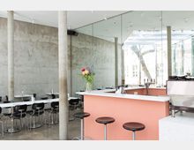 Helles Café mit moderner Einrichtung, bestehend aus einer Theke mit rosa Verkleidung, schwarzen Barhockern und Tischen mit schwarzen Stühlen im Hintergrund. Die Wände sind aus Sichtbeton, und eine Glasfront bietet Blick auf einen Baum außerhalb des Gebäudes.
