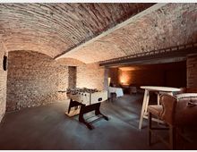 Gewölbekeller mit freiliegenden Backsteinwänden und gewölbter Decke, eingerichtet mit einem Kickertisch, hohen Holztischen und gepolsterten Stühlen. Im Hintergrund befinden sich weitere Sitzgelegenheiten und gedämpftes Licht.