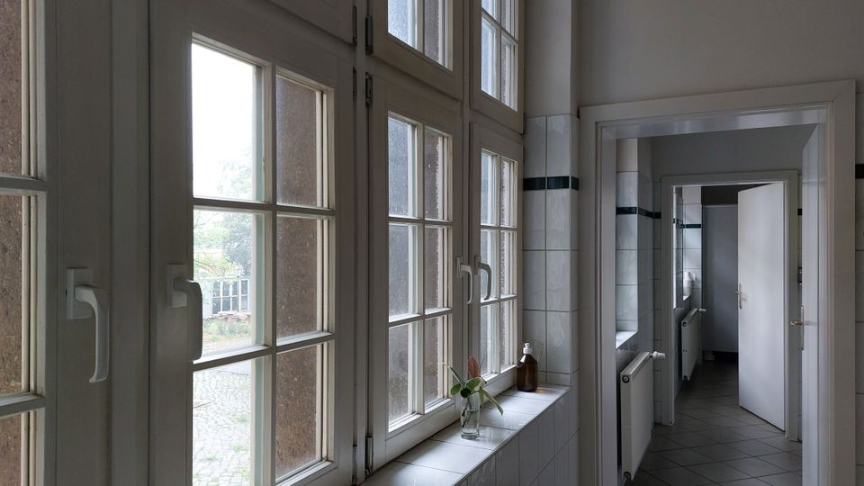 Ein Flur in einem Gebäude mit weißen Fliesen an den Wänden, großen Fenstern mit Sprossenrahmen auf der linken Seite und weißen Türen, die zu angrenzenden Räumen führen. Auf der Fensterbank steht eine kleine Vase mit Blumen und eine braune Flasche.