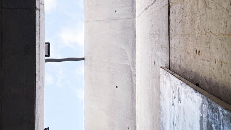 Beton-Innenraum mit hohen, glatten Wänden, einem großen rechteckigen Deckenfenster, das den Himmel zeigt, und Metallstreben im oberen Bereich. Auf einer der Wände ist ein abstraktes Kunstwerk sichtbar.