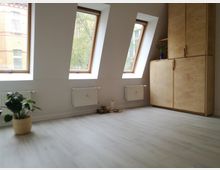 Heller Raum mit drei großen Dachfenstern, durch die Tageslicht einfällt. Der Boden ist mit hellem Holzlaminat ausgelegt, an der rechten Wand steht ein hoher Schrank aus naturbelassenem Holz, und eine Zimmerpflanze in einem Korb befindet sich links im Vordergrund.