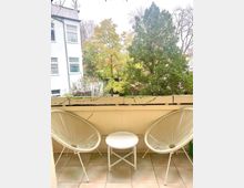 Ein kleiner Balkon mit zwei weißen Stühlen und einem runden weißen Beistelltisch auf hell gefliesten Boden. Im Hintergrund sind grüne Bäume und ein benachbartes Wohnhaus zu sehen.